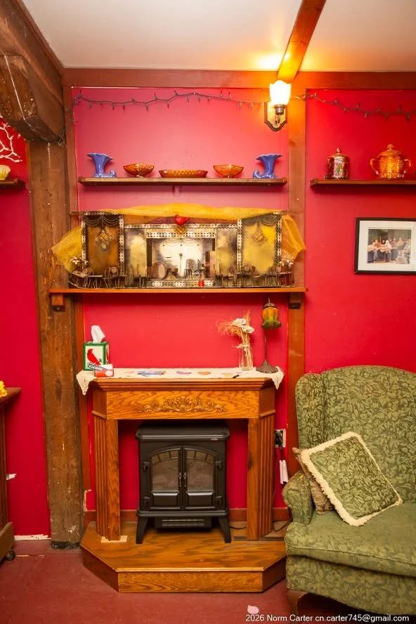 A fireside corner: green wingback armchair beside the wood-mantel fireplace, shelves of carnival glass above, red wall behind.